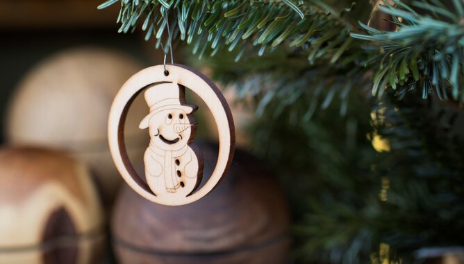 BIO Christbäume | © SONNENTOR Auf dem Foto sieht man einen Christbaumaufhänger aus Holz mit einem Schneemannmotiv. Das Dekostück hängt auf einer Tanne. | © SONNENTOR